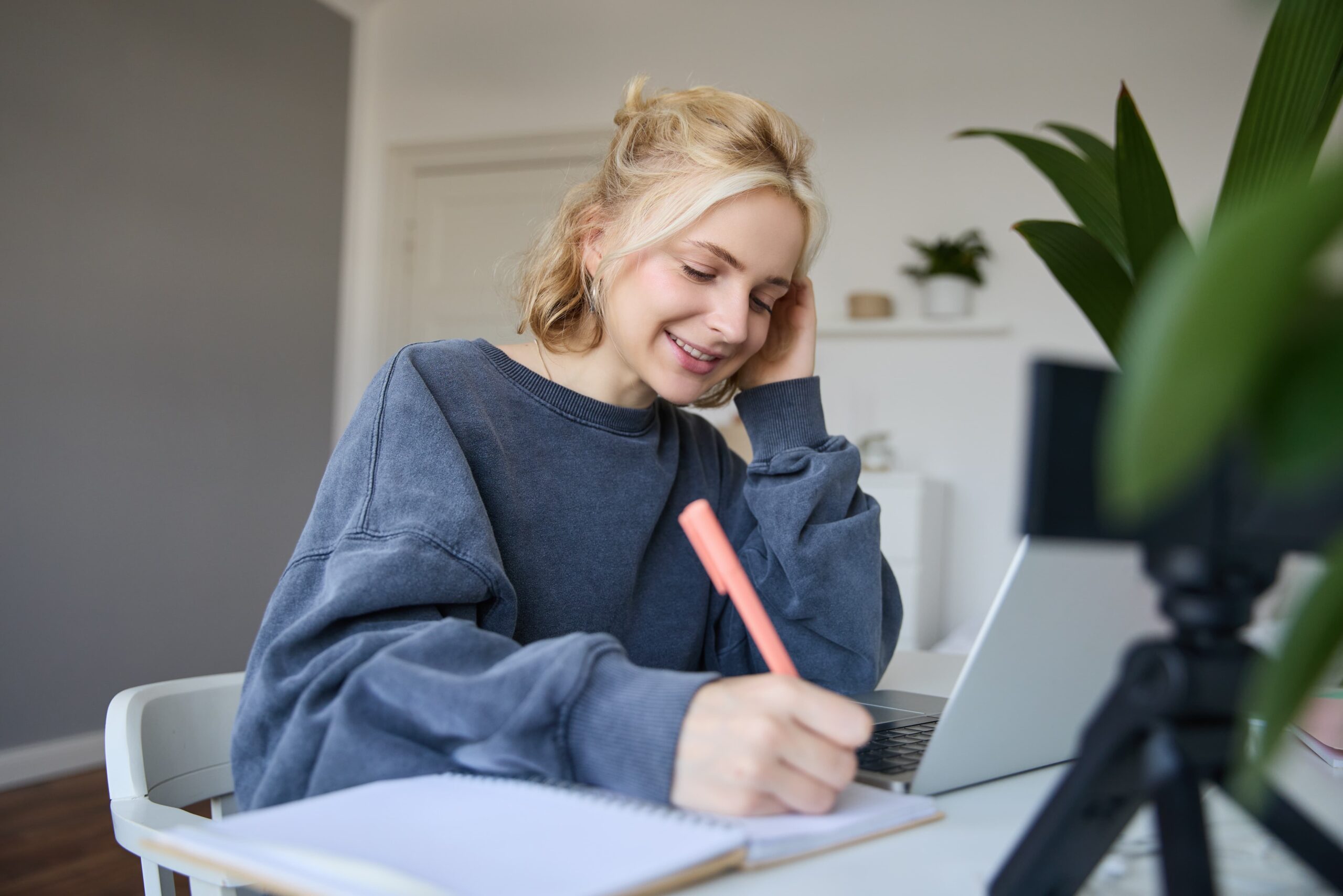 Kvinne som skriver på en notatblokk foran en laptop, smilende og fokusert, som en del av arbeidet med å lage engasjerende tekster for arrangementer.